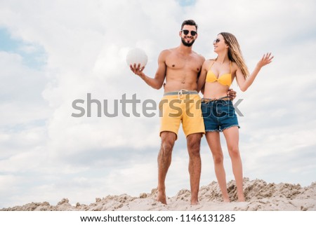 beautiful young couple with volleyball ball standing on sandy dune in front of cloudy sky
