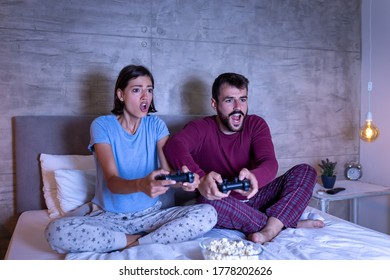 Beautiful Young Couple Wearing Pajamas, Having Fun At Home, Playing Video Games In Bed At Night, Woman Excited After Winning The Game, Man Disappointed After Losing