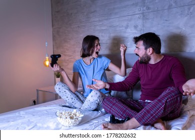 Beautiful Young Couple Wearing Pajamas, Having Fun At Home, Playing Video Games In Bed At Night, Woman Excited After Winning The Game, Man Disappointed After Losing