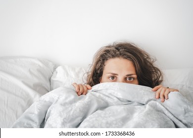 Beautiful Young Curly Woman Lies On The Bed And Looks Out From Under The Covers, Looking Seriously At The Camera