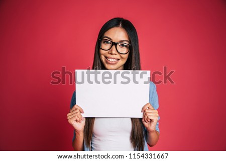 Beautiful young cute lovely asian woman in glasses with white blank board or banner in hands for advertisement over pink background