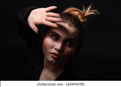 Beautiful Young Dark-haired Girl With Brown Eyes In Dark Clothes. Low Key Portrait On A Dark Background. The Face Of The Model Is Lit By Bright Light. Close-up Portrait Of A Hand In A Frame.