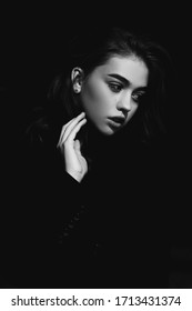 Beautiful Young Dark-haired Girl With Brown Eyes In Dark Clothes. Low Key Portrait On A Dark Background. The Face Of The Model Is Lit By Bright Light. Close-up Portrait Of A Hand In A Frame.