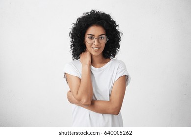 Beautiful Young Dark-skinned Female Wearing Stylish Round Glasses And Black Casual T-shirt, Making Awkward Gesture, Touching Her Neck During Intense Conversation With Someone, Trying To Smile