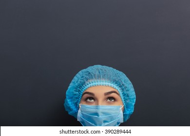 Beautiful young doctor in white medical gown, cap and mask is looking up, standing against blackboard, close-up