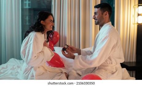 Beautiful And Young Ethnic Couple Spending Valentines Day Together In A Fancy Hotel Suite Room. Handsome Man Asks The Pretty Female To Marry Him, Woman Looks Shocked And Happy,Surrounded With Balloons