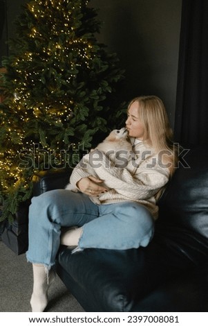 A beautiful young European girl with her Pomeranian cream-white spitz dog is sitting on the sofa, behind a Christmas tree with lights and a Christmas atmosphere