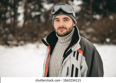 Beautiful young European man in a gray sweater and ski jacket, hat, goggles, ski orange boots in the forest in winter. Beautiful portrait, ski resort, vacation, mountain, active sport