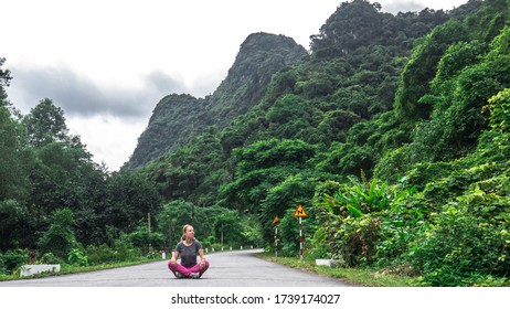 A Beautiful Young European Woman Is Sitting In The Lotus Pose On The Road Surrounded By Saturated Green Trees In The Middle Of Cat Ba Island In Northern Vietnam

Concept: Traveling