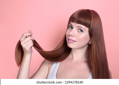 Beautiful young fair-skinned woman holding a curl of hair.  She has smooth long healthy hair and thick bangs. Face of model close-up on pink background. Hair care concept.