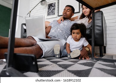 Beautiful Young Family African American Dad Asian Mom And Charming Little Daughter Watching Tv Shows And Cartoons Using Laptop Smartphone And Wi Fi Internet Sitting On Floor In Modern Interior