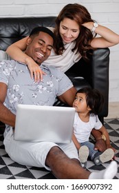 Beautiful Young Family African American Dad Asian Mom And Charming Little Daughter Watching Tv Shows And Cartoons Using Laptop Smartphone And Wi Fi Internet Sitting On Floor In Modern Interior