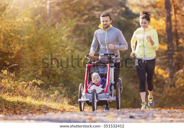 Beautiful young family with baby in jogging\
stroller running outside in autumn\
nature