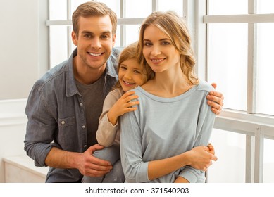 Beautiful young family looking in camera, smiling and hugging while sitting near the window at home