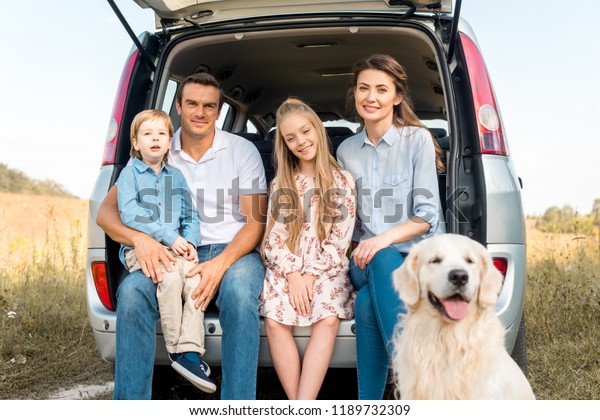 beautiful young family with retriever dog\
sitting in car trunk and looking at camera in\
field