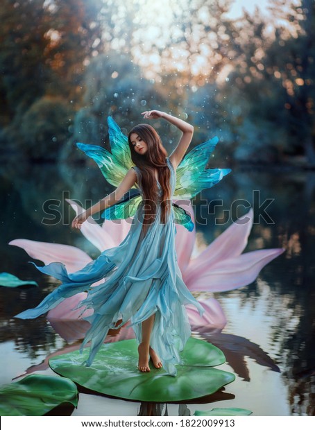 Beautiful young fantasy woman in image river fairy\
dances on water pink lily flower. long silk dress flies in wind\
motion butterfly wings magic shiny. Art Girl pixie. Background dark\
nature, blue lake