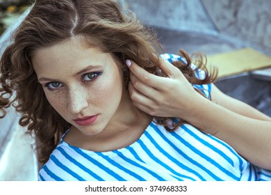 Beautiful young fashion model with freckles on her face and light blue striped dress and fashion makeup and hairstyle is lying down in metalic silver boat on lake, posing and looking at camera.