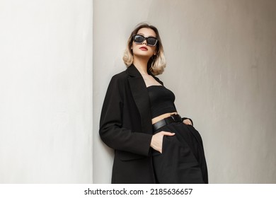 Beautiful young fashionable businesswoman with fashionable sunglasses wearing a fashionable black elegant blazer and a stylish top with pants is standing near the white wall on the street - Powered by Shutterstock