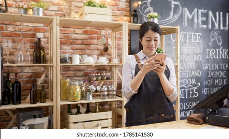 beautiful young female barista in apron is using smartphone and smiling while standing in bar counter at cafe store. elegant lady staff of coffeehouse relax in workplace chatting online on cellphone. - Powered by Shutterstock