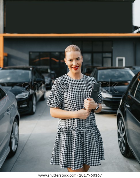 Beautiful young female car dealer standing in\
front of parked cars. Used car dealership concept. She is standing,\
posing and looking at\
camera.