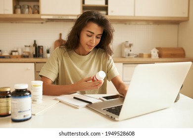 Beautiful Young Female Dietician Working From Home, Using Portable Computer, Typing Article About Vitamins, Holding Bottle Of Food Supplements, Reading Label On It. Health, Wellness And Dieting