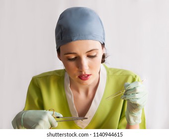 Beautiful Young Female Doctor Wearing Green Surgical Scrubs, Blue Scrub Cap With Surgical Gloves, Placing Sutures In Front Of A White Background