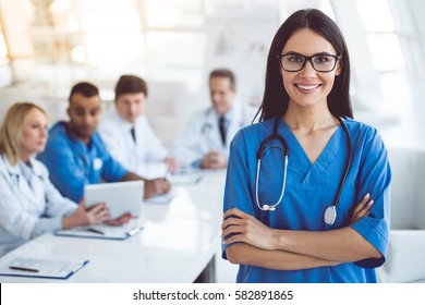 Beautiful young female medical doctor is looking at camera and smiling while her colleagues are sitting in the background - Powered by Shutterstock