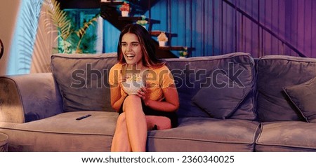 Beautiful young female sitting on sofa watching comedy movie eating popcorn snack enjoy weekend at indoor home. Happy Indian woman laughing looking funny web series on television spend free time
