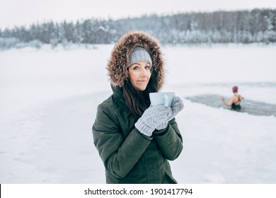 Beautiful young female smiling and drinking tea in nature Winter swimming. Woman in frozen lake ice hole. Swimmers wellness in icy water. How to swim in cold water Gray hat and gloves swimming clothes