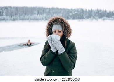 Beautiful Young Female Smiling And Drinking Tea In Nature Winter Swimming. Woman In Frozen Lake Ice Hole. Swimmers Wellness In Icy Water. How To Swim In Cold Water Gray Hat And Gloves Swimming Clothes