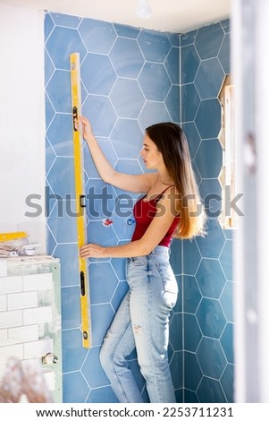 Beautiful young female tiler laying textured hexagon tiles on bathroom wall, checking level with tool at construction site