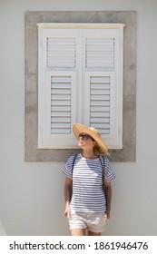 Beautiful young female tourist woman wearing sun hat, standing and relaxing in front of vinatage wooden window in old Mediterranean town while sightseeing on hot summer day.