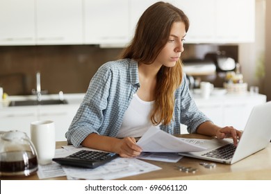 Beautiful young female wearing casual shirt calculating family budget, looking through expenses while managing home accounts, paying utility bills online using wifi on generic notebook computer