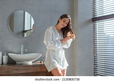 Beautiful Young Female In White Silk Robe Brushing Her Hair With Comb While Standing In Modern Loft Bathroom Interior, Happy Millennial Woman Making Daily Beauty Routine At Home, Copy Space