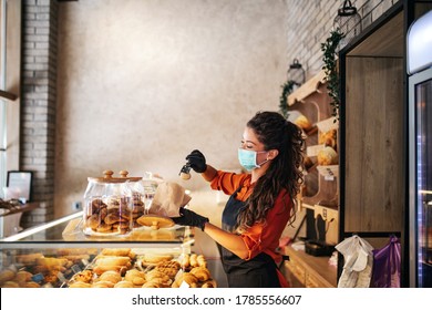 Beautiful young female worker with protective mask on face working in bakery.