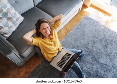 Beautiful young female in yellow shirt sitting on the floor next to the sofa on the carpet with laptop, freelancer and home office concept.
