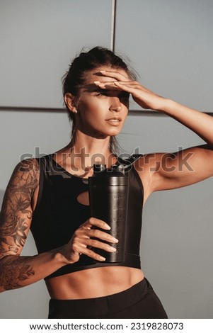 Beautiful young fit tanned caucasian woman with tattoo holding a protein shaker or water bottle on grey wall background on summer sunny day