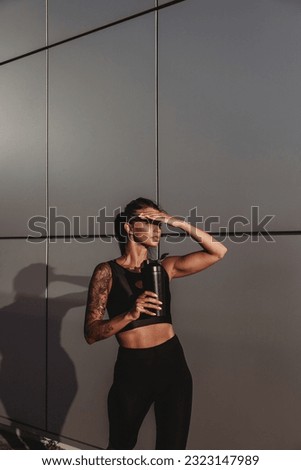Beautiful young fit tanned caucasian woman with tattoo holding a protein shaker or water bottle on grey wall background on sunset