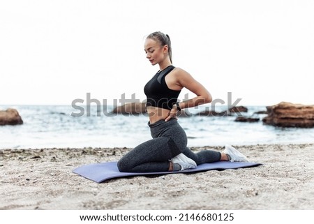 Beautiful young fit woman in sportswear practicing stretching on yoga mat alone on wild beach. Healthy lifestyle concept