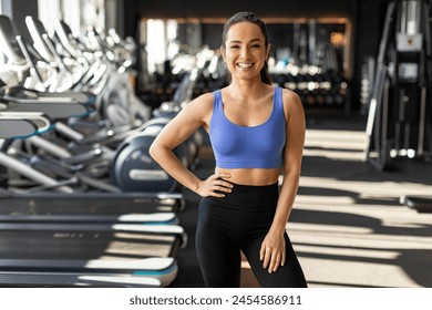 Beautiful young fitness lady in sportswear with hand on hip standing in sports gym, posing and smiling at camera during workout - Powered by Shutterstock