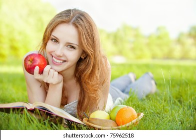 A beautiful young girl with an apple lying on the grass and reading a book beside a basket of fruit is, on a background of green nature - Powered by Shutterstock