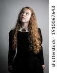 Beautiful young girl in black 1890s English Victorian 18th century child period dress with elegant white lace collar antique broach jewelery and long curly pretty hair looking up to sky