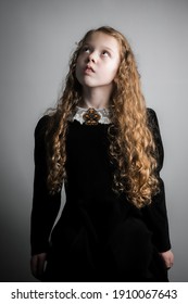 Beautiful young girl in black 1890s English Victorian 18th century child period dress with elegant white lace collar antique broach jewelery and long curly pretty hair looking up to sky