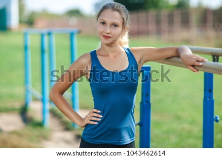 Beautiful young girl in a blue shirt and leggings on the uneven bars on the outdoor sports ground in summer looking at camera and smiling