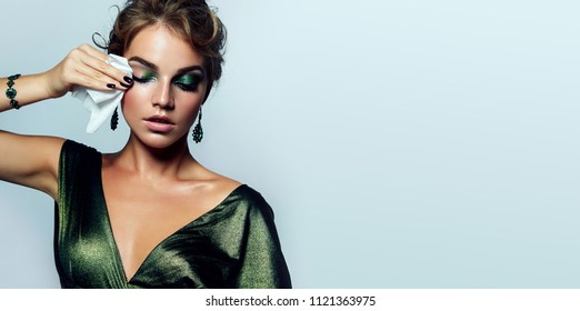 beautiful young girl with a bright make-up and in a shiny green dress striatet makeup from her face with a wet napkin. Hairstyle - curls are gathered in a bun.fashion, beauty, makeup, cosmetics. - Shutterstock ID 1121363975