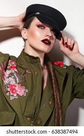 Beautiful Young Girl With Bright Make-up In A Green Trench Coat And A Black Cap Stands In The Studio On A White Background. Military Uniform, Army, Fashion, Beauty, Hairstyle - Fish Tail. Clothing.