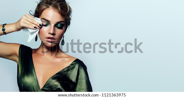 beautiful young girl with a bright make-up and\
in a shiny green dress striatet makeup from her face with a wet\
napkin. Hairstyle - curls are gathered in a bun.fashion, beauty,\
makeup, cosmetics.