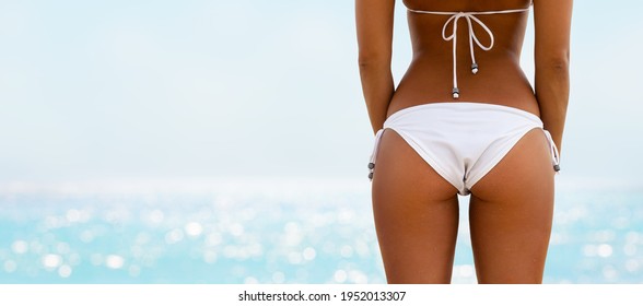 Beautiful Young Girl With Bronze Tan On The Beach In A White Bikini. Close-up Of Beautiful Woman With Sexy Booty In A Swimsuit Posing On The Beach. Summer Beach Concept.