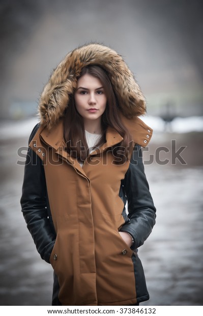 Beautiful young girl with brown coat trimmed with\
gray fur enjoying the winter scenery in park. Teenage girl with\
black leather sleeves coat posing in winter concept. Young female\
bright cold day