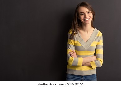 Beautiful Young Girl In Casual Clothes Is Looking At Camera And Smiling, Standing With Crossed Arms Against Dark Background